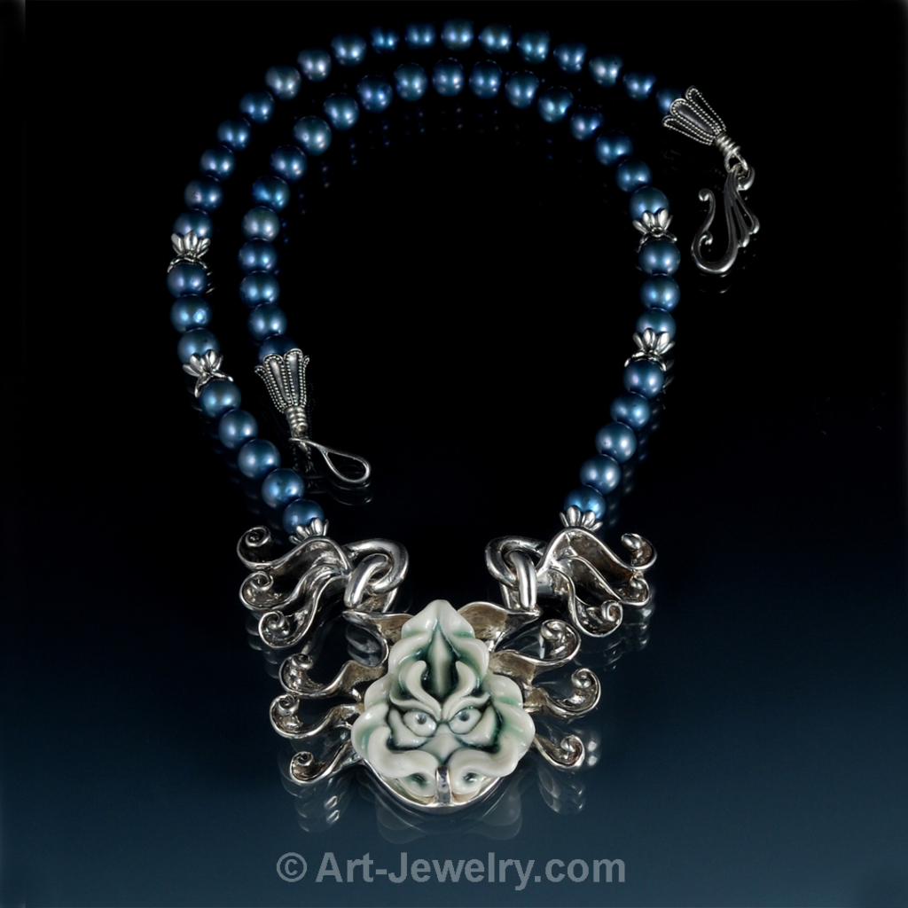 Hand‑carved porcelain dragon face with blue‑green celadon glaze set in a custom sterling silver bezel with radiating spiral wings, strung on raven‑black pearls.